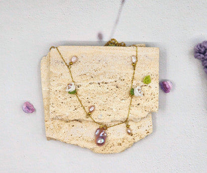 Handcfrafted vintage-style necklace featuring a centrepiece purple-pink metallic Baroque pearl, flanked by pink baroque pearls and micro crochet white lily of the valley blooms. 18K gold-plated titanium stell chain.